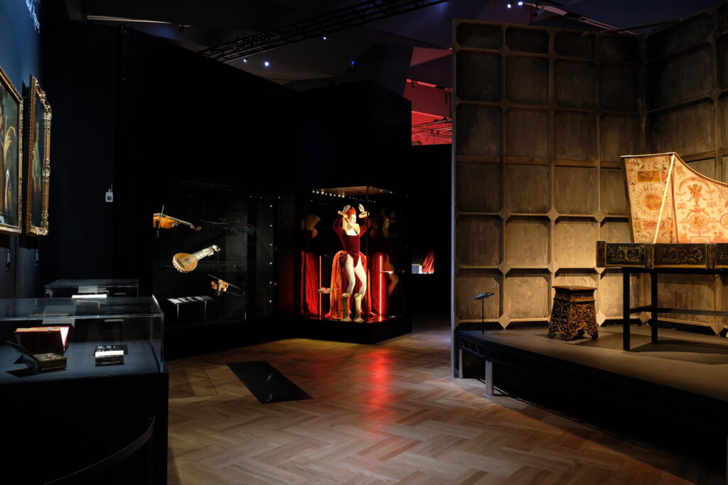 Dark lit exhibition space with a piano on the stage at the V&A